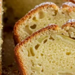 Gros plan d'un gâteau aux poires et pommes fraîchement coupé, montrant les tranches moelleuses avec des morceaux de fruits visibles, saupoudrées de sucre glace. La croûte est dorée et croustillante, créant un contraste appétissant avec l'intérieur tendre.