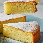 Tranches de gâteau yaourt sans gluten moelleux saupoudrées de sucre glace, présentées sur une assiette bleu clair. Une tranche est soulevée par une spatule dorée.