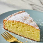 Une tranche de gâteau yaourt sans gluten, saupoudrée de sucre glace, est présentée sur une assiette bleu clair avec une fourchette dorée à côté. La tranche de gâteau a une texture moelleuse et une couleur dorée.