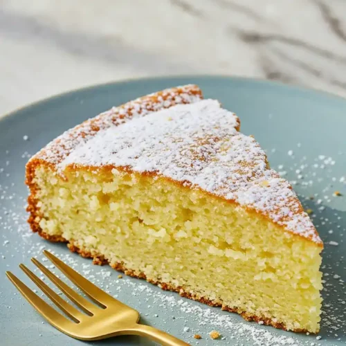 Une tranche de gâteau yaourt sans gluten, saupoudrée de sucre glace, est présentée sur une assiette bleu clair avec une fourchette dorée à côté. La tranche de gâteau a une texture moelleuse et une couleur dorée.