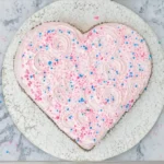 Vue de dessus d'un gâteau Saint-Valentin en forme de cœur, recouvert d'un glaçage rose et de décorations colorées.