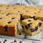 Un appétissant gateau gouter aux myrtilles, doré et moelleux, est présenté sur une grille. Une portion a été coupée, révélant une mie aérée parsemée de myrtilles juteuses. Quelques pépites de chocolat sont dispersées autour, ajoutant une touche gourmande à ce dessert simple et parfait pour une collation réconfortante.