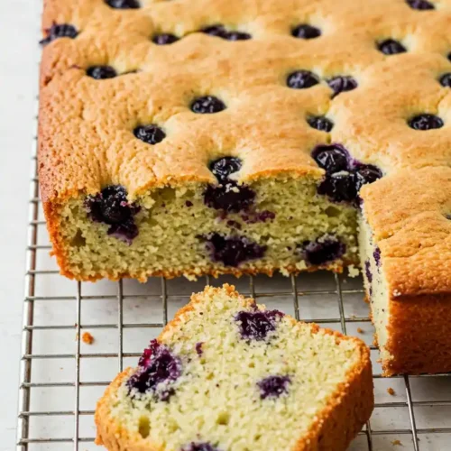 Une vue rapprochée d'un délicieux gateau gouter aux myrtilles, fraîchement sorti du four et posé sur une grille. Une part a été coupée, révélant une texture moelleuse et aérée, parsemée de myrtilles éclatantes. Ce gateau gouter simple et savoureux est une gourmandise parfaite pour accompagner un café ou un thé, idéal pour une pause gourmande.
