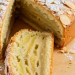 Une tranche de gateau invisible aux pommes​, décorée d'amandes effilées et saupoudrée de sucre glace, est délicatement séparée du reste du gâteau, exposant les couches de pommes moelleuses sur une planche de bois.