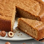 Un appétissant gâteau noisette rond, tranché, présenté sur une assiette blanche avec quelques noisettes entières. Une part est soulevée avec une spatule, révélant une texture moelleuse et une couleur dorée irrésistible.
