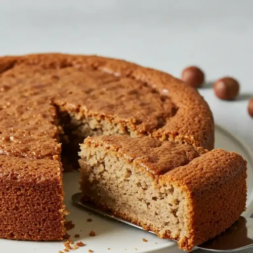 Vue en plongée d'un délicieux gâteau noisette, une tranche en étant délicatement prélevée à l'aide d'une spatule. Des noisettes entières sont éparpillées autour de l'assiette, soulignant la saveur principale de ce dessert gourmand.