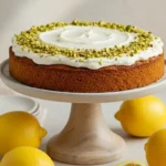 Voici une photo appétissante d'un gateau au semoule, probablement une recette gateau au semoule au citron, décoré avec un glaçage blanc crémeux et des pistaches hachées, présenté sur un élégant présentoir à gâteau entouré de citrons frais.