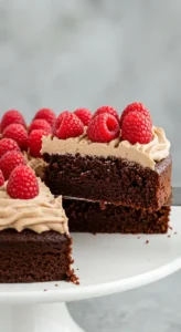 Un délicieux Gâteau Chocolat Sans Farine, nappé d'une crème légère et garni de framboises fraîches, dont une part est en train d'être coupée.