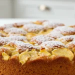 Un délicieux gâteau léger aux pommes, parsemé de sucre glace, présenté sur une assiette blanche. Sa texture moelleuse et ses morceaux de pommes juteuses en font un dessert parfait pour toutes les occasions.