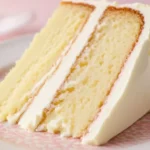 Une part appétissante de gateau sans oeuf à étages, avec des couches de biscuit jaune pâle et un glaçage blanc onctueux, servie sur une assiette à motifs roses.
