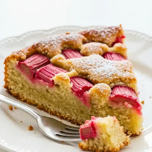 Une part appétissante de gâteau rhubarbe saupoudrée de sucre glace, servie sur une assiette blanche avec une fourchette, parfaite pour une dégustation acidulée et gourmande.