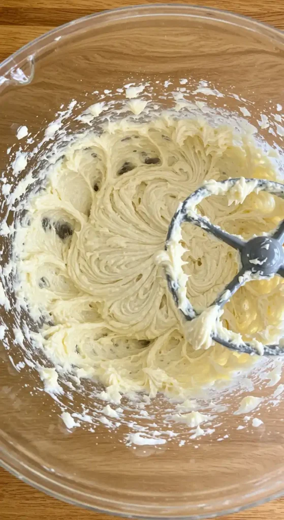Un bol en verre transparent rempli d'une crème onctueuse et jaune pâle, en cours de mélange avec un batteur électrique gris. La crème a une texture lisse et uniforme, parfaite pour réaliser un glaçage de gâteau anniversaire facile et délicieux.