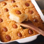 Portion de gateau arabe : Un gateau arabe fraîchement cuit et décoré d'amandes est présenté dans un plat, avec une portion prélevée à l'aide d'une spatule en bois, révélant sa texture moelleuse et dorée.