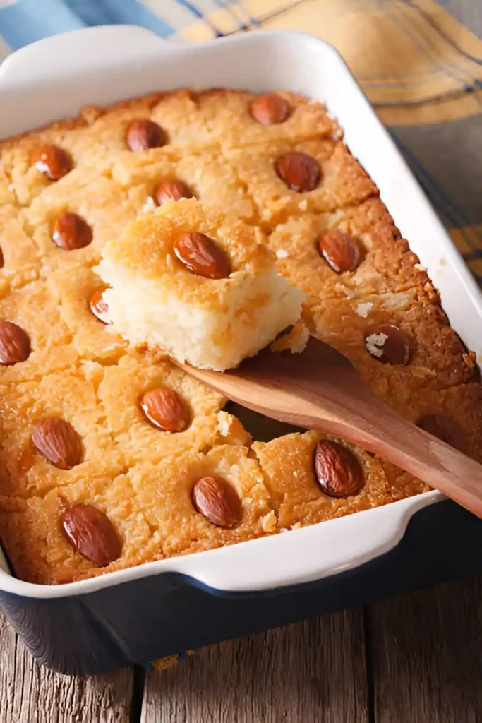 Portion de gateau arabe : Un gateau arabe fraîchement cuit et décoré d'amandes est présenté dans un plat, avec une portion prélevée à l'aide d'une spatule en bois, révélant sa texture moelleuse et dorée.