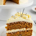 Gros plan d'une part de gâteau moelleux, décorée avec une crème blanche onctueuse et des billes dorées et argentées. Deux pics argentés ajoutent une touche festive. La part est dressée sur une assiette blanche, devant un reste de gâteau possiblement un gâteau patate douce, présenté sur un présentoir.