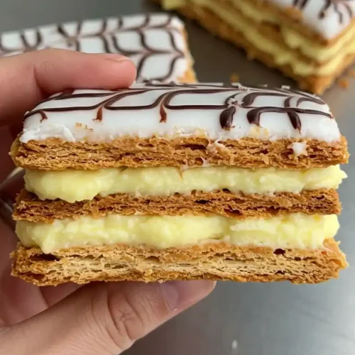 Un millefeuille facile et rapide, superbement réalisé, présenté dans une main. On admire les couches de pâte feuilletée croustillantes et dorées, alternant avec une généreuse garniture de crème pâtissière à la vanille. Le glaçage blanc, décoré de motifs délicats au chocolat, couronne ce dessert classique, parfait pour une dégustation gourmande.