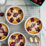 Vue de dessus de plusieurs ramequins de dessert cerise cuits, garnis d'amandes effilées et de sucre glace, disposés sur une grille avec des cerises fraîches, des amandes supplémentaires, du sucre glace et des cuillères vintage à proximité, évoquant une ambiance gourmande et raffinée.