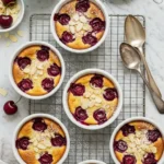 Présentation artistique de ramequins de dessert cerise, saupoudrés d'amandes effilées et de sucre glace, sur une grille, avec des cerises fraîches, des amandes, un tamis à sucre glace et des cuillères argentées, capturant l'essence d'une pâtisserie maison élégante.