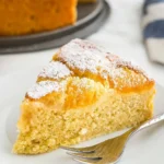 Gros plan d'une part de gateau abricot frais, délicatement saupoudrée de sucre glace, présentée sur une assiette blanche avec une fourchette. La texture moelleuse du gâteau contraste avec la douceur des abricots.
