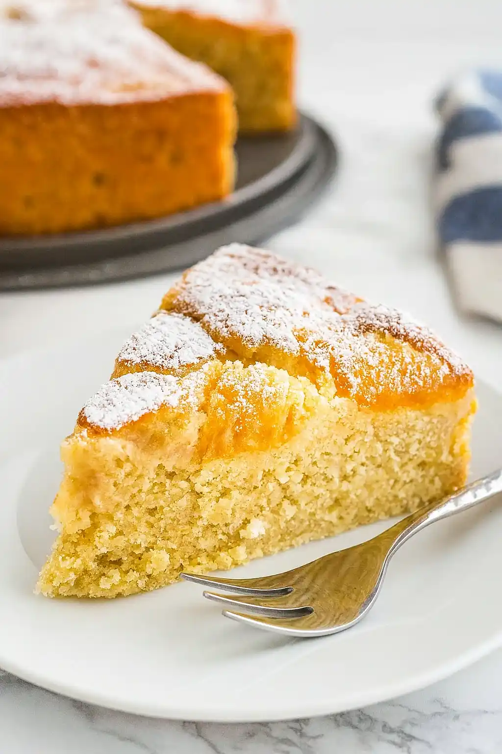 Gros plan d'une part de gateau abricot frais, délicatement saupoudrée de sucre glace, présentée sur une assiette blanche avec une fourchette. La texture moelleuse du gâteau contraste avec la douceur des abricots.
