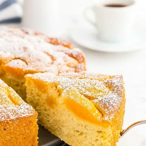 Une part de gateau abricot frais saupoudrée de sucre glace, servie avec une tasse de café. Le gâteau moelleux révèle des morceaux d'abricots juteux.