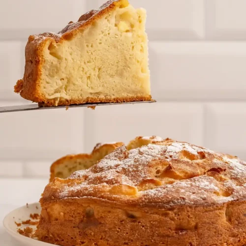 Une tranche du gâteau moelleux aux pommes de ma grand-mère est délicatement soulevée avec une spatule, révélant sa texture aérée et son parfum irrésistible, tandis que le reste du gâteau repose sur une assiette, prêt à être dégusté.