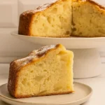 Une part moelleuse de gâteau aux pommes, saupoudrée de sucre glace, est présentée sur une assiette, avec le reste du gâteau en arrière-plan, évoquant les souvenirs du gâteau moelleux aux pommes de ma grand-mère.