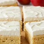 Gros plan savoureux de carrés de Gâteau nuage aux pommes, chacun recouvert d'une épaisse couche de glaçage blanc crémeux. La texture moelleuse de la mie est visible, et des pommes fraîches, rouges et appétissantes, trônent en arrière-plan, promettant une saveur fruitée et automnale. Parfait pour une pause gourmande !