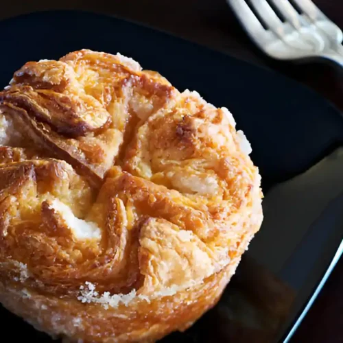 Un délicieux kouign amann individuel, doré et caramélisé, présenté sur une assiette noire brillante, accompagné d'une fourchette. La pâtisserie bretonne révèle ses couches feuilletées et son aspect croustillant, promettant une dégustation gourmande.
