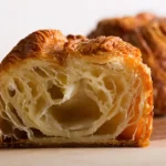 Gros plan d'un kouign amann coupé en deux, révélant la texture feuilletée et aérée de la pâte. L'intérieur est clair et beurré, tandis que l'extérieur est doré et croustillant, typique de cette spécialité bretonne, faisant ressortir le côté gourmand du kouign amann. Un autre kouign amann flou est visible en arrière plan.