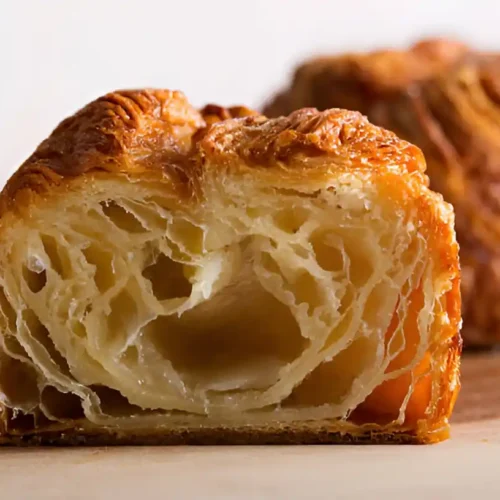 Gros plan d'un kouign amann coupé en deux, révélant la texture feuilletée et aérée de la pâte. L'intérieur est clair et beurré, tandis que l'extérieur est doré et croustillant, typique de cette spécialité bretonne, faisant ressortir le côté gourmand du kouign amann. Un autre kouign amann flou est visible en arrière plan.