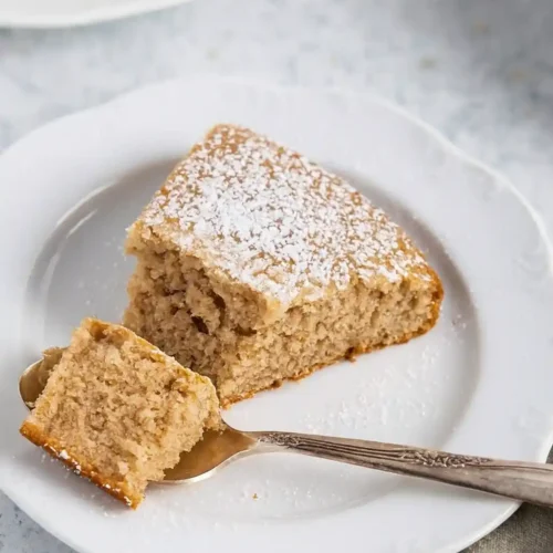 Gateau creme de marron : Une part de gâteau moelleux saupoudrée de sucre glace, présentée sur une assiette blanche avec une cuillère contenant une bouchée.