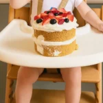 Un adorable bébé en chaise haute, prêt à déguster son gateau 1 an, garni de crème et de fruits frais comme des fraises et des myrtilles.