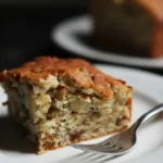 Un morceau de gâteau flocon d'avoine doré, servi sur une assiette blanche avec une fourchette. La texture du gâteau est visible, avec une croûte légèrement croustillante et un intérieur moelleux. Un autre morceau de gâteau est flou en arrière-plan.