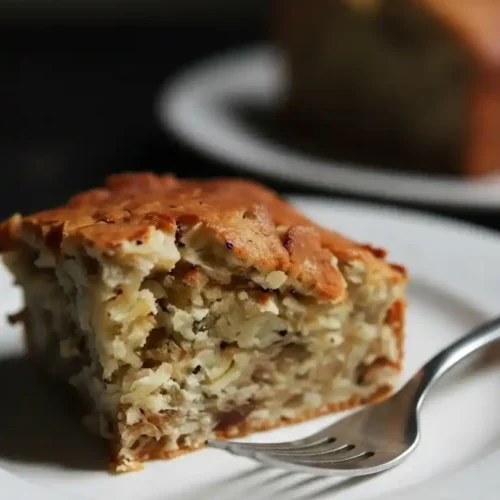 Un morceau de gâteau flocon d'avoine doré, servi sur une assiette blanche avec une fourchette. La texture du gâteau est visible, avec une croûte légèrement croustillante et un intérieur moelleux. Un autre morceau de gâteau est flou en arrière-plan.