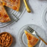 Une vue plongeante appétissante d'une table dressée avec un délicieux gâteau aux mirabelles. Un morceau de gâteau repose sur une assiette en verre délicate, prêt à être dégusté avec une fourchette. À côté, on aperçoit le reste du gâteau aux mirabelles sur une grille, avec un couteau doré élégant. Des bols remplis d'amandes et de mirabelles fraîches complètent cette scène gourmande.