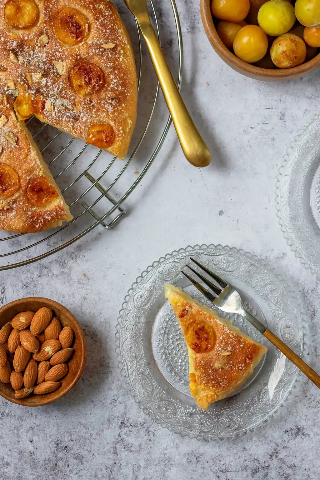 Une vue plongeante appétissante d'une table dressée avec un délicieux gâteau aux mirabelles. Un morceau de gâteau repose sur une assiette en verre délicate, prêt à être dégusté avec une fourchette. À côté, on aperçoit le reste du gâteau aux mirabelles sur une grille, avec un couteau doré élégant. Des bols remplis d'amandes et de mirabelles fraîches complètent cette scène gourmande.