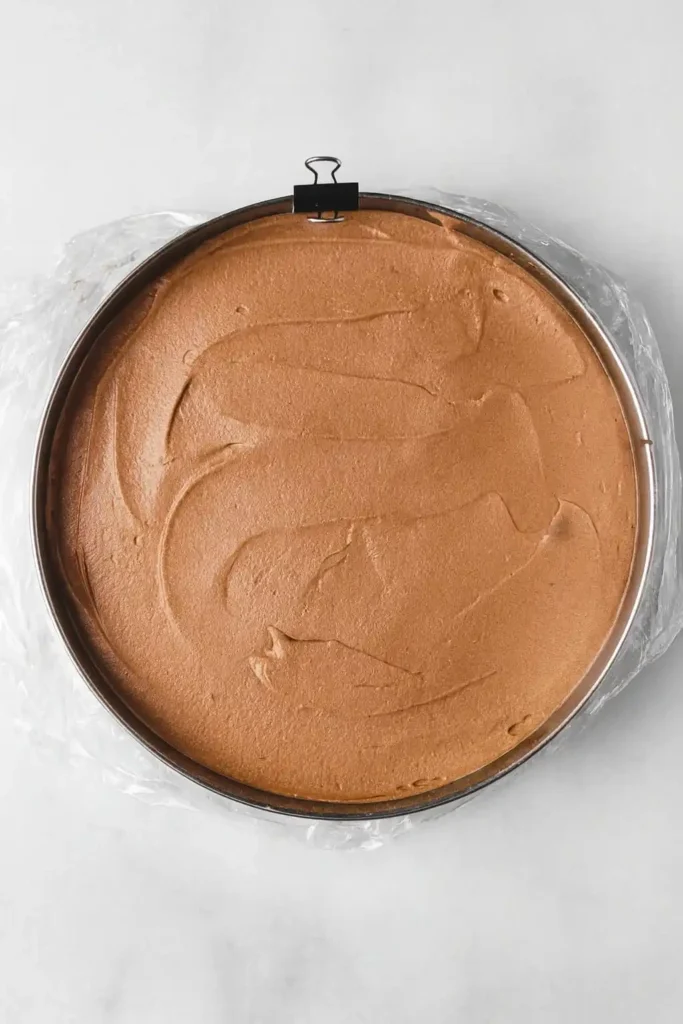 Un moule à charnière rond contient une mousse au chocolat lisse et uniformément étalée, prête à être réfrigérée ou cuite. Le moule est doublé de film plastique.