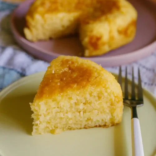 Un morceau de gâteau jaune, ressemblant à un gâteau de riz avec une surface brillante, est présenté sur une assiette verte à côté d'une fourchette. En arrière-plan, on distingue le reste du gâteau sur une autre assiette. Cette présentation soignée pourrait évoquer un gâteau peu calorique, mais sans information supplémentaire, son origine industrielle reste incertaine.