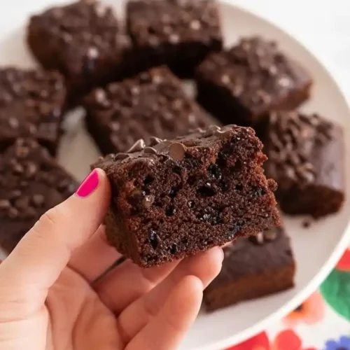 Une main tenant une part de gâteau au chocolat moelleux, avec un plateau rempli d'autres parts en arrière-plan, une idée gourmande pour un gâteau goûter enfant.