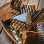 Délicieux gâteau russe multicouche avec glaçage au chocolat, servi avec des cuillères décoratives sur un plateau en bois rustique.