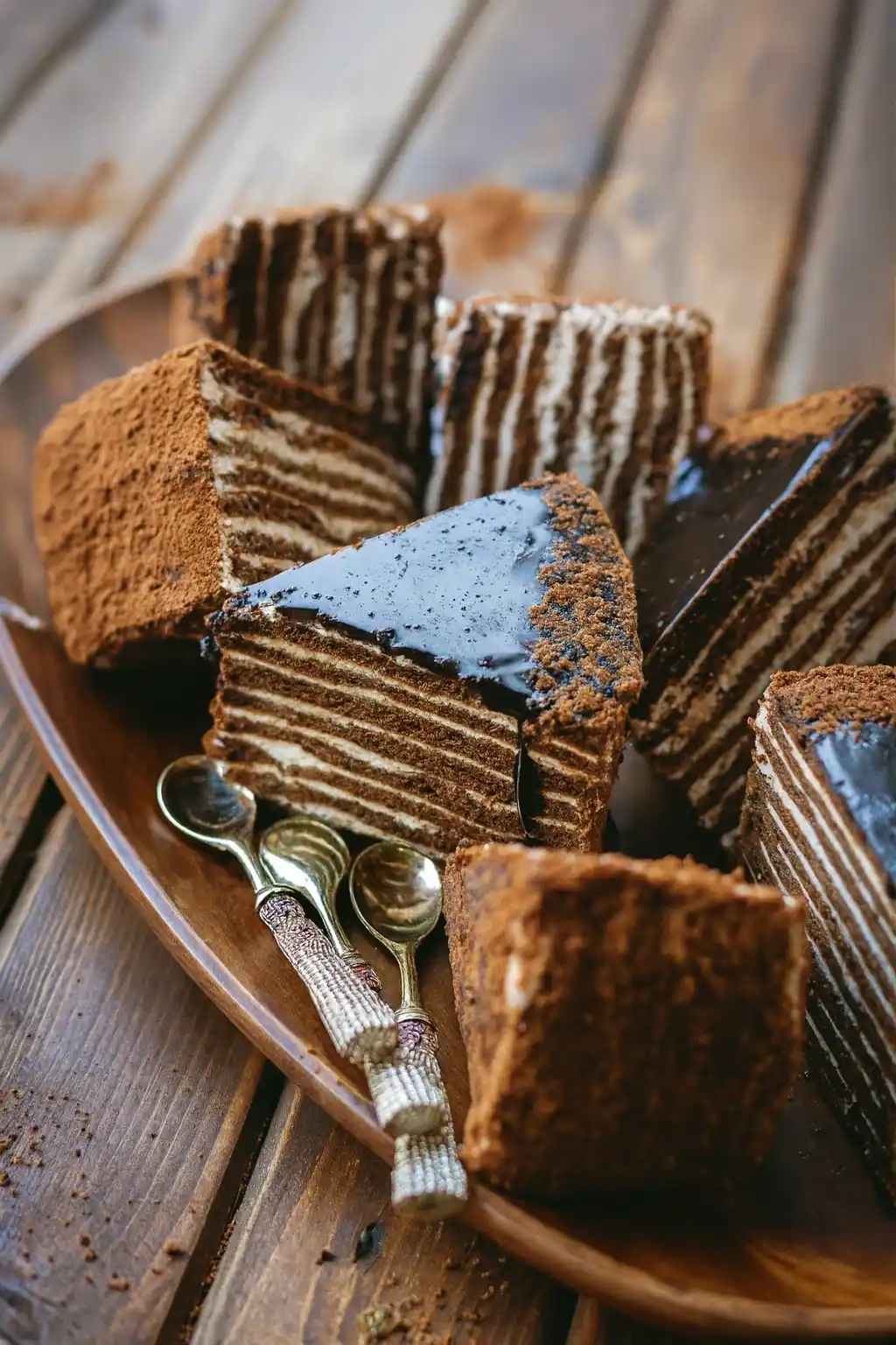 Délicieux gâteau russe multicouche avec glaçage au chocolat, servi avec des cuillères décoratives sur un plateau en bois rustique.