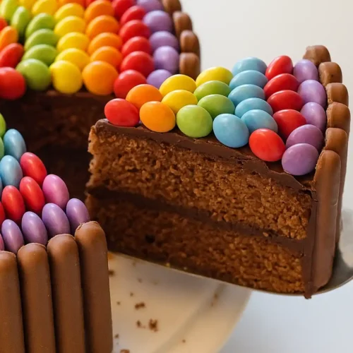 Un délicieux gâteau Smarties est mis en évidence. Une tranche est soulevée, révélant un intérieur chocolaté avec un remplissage crémeux. Le dessus du gâteau est décoré d'une rangée colorée de Smarties disposés en arc-en-ciel, tandis que les côtés sont ornés de bâtonnets de chocolat.
