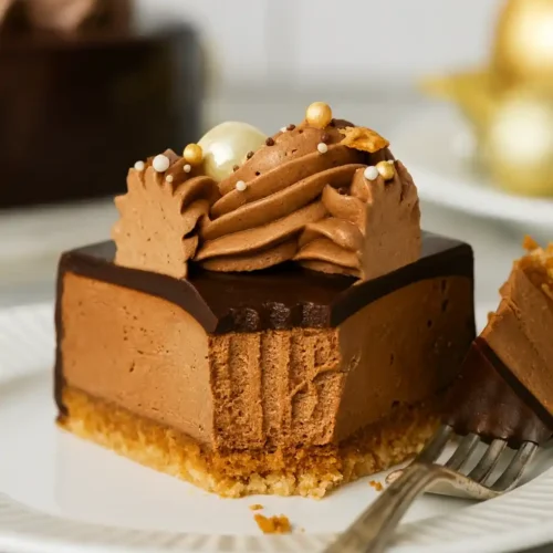 Un délicieux morceau de gâteau Trianon est présenté sur une assiette blanche, avec une fourchette à proximité. On peut voir les différentes couches : un biscuit croustillant, une mousse au chocolat onctueuse, un glaçage brillant et une décoration de crème au chocolat. Un autre gâteau est visible en arrière-plan.
