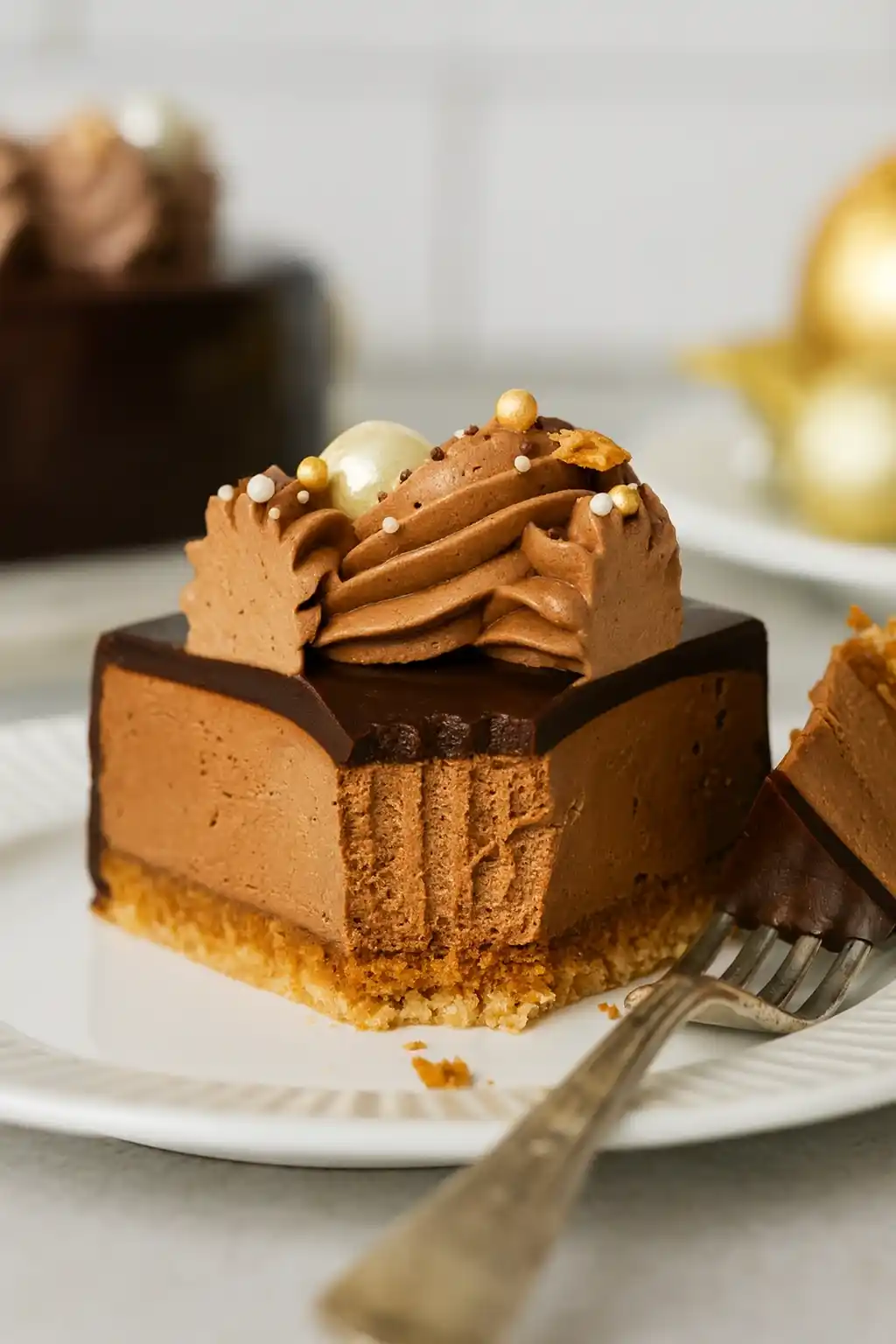 Un délicieux morceau de gâteau Trianon est présenté sur une assiette blanche, avec une fourchette à proximité. On peut voir les différentes couches : un biscuit croustillant, une mousse au chocolat onctueuse, un glaçage brillant et une décoration de crème au chocolat. Un autre gâteau est visible en arrière-plan.