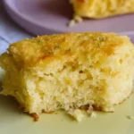 Un morceau de gâteau jaune, avec une bouchée manquante, est présenté sur une assiette vert clair, décoré de quelques herbes vertes. Un autre morceau de ce qui semble être le même gâteau est visible sur une assiette violette en arrière-plan. L'apparence légère et aérée du gâteau pourrait suggérer une option peu calorique, mais l'absence d'informations empêche de confirmer si c'est un gâteau industriel ou fait maison.