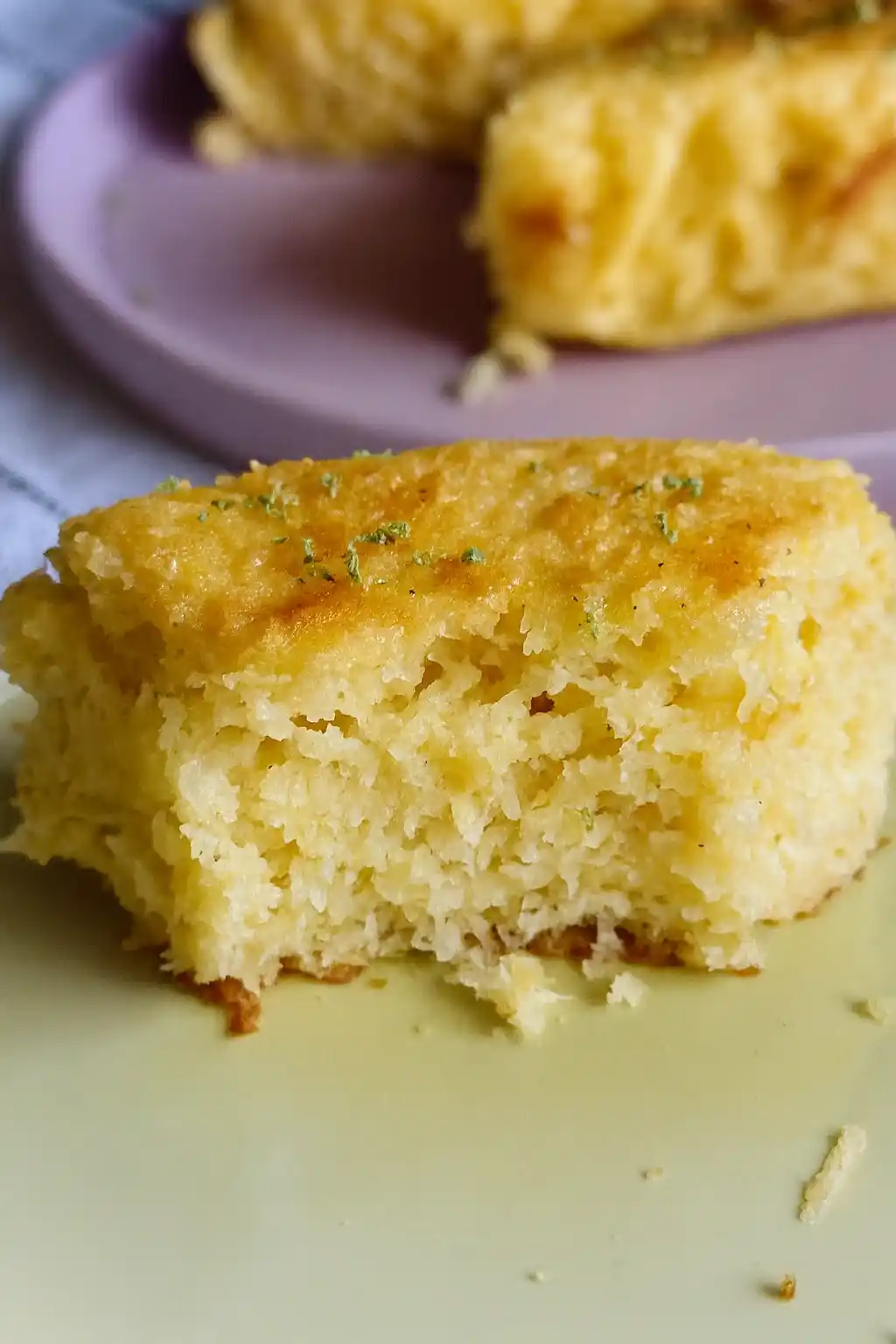 Un morceau de gâteau jaune, avec une bouchée manquante, est présenté sur une assiette vert clair, décoré de quelques herbes vertes. Un autre morceau de ce qui semble être le même gâteau est visible sur une assiette violette en arrière-plan. L'apparence légère et aérée du gâteau pourrait suggérer une option peu calorique, mais l'absence d'informations empêche de confirmer si c'est un gâteau industriel ou fait maison.