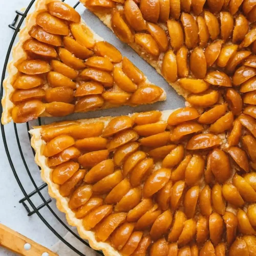 Une délicieuse tarte aux mirabelles, dont une part a été coupée, repose sur une grille. Les mirabelles sont disposées en rangées régulières et brillent grâce au glaçage. Une illustration parfaite de la tarte mirabelle recette.