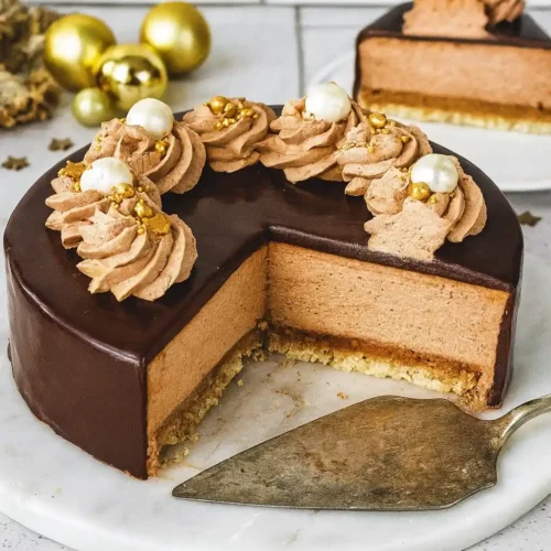 Un somptueux gâteau Trianon rond, orné de crème au chocolat, de perles décoratives et d'un glaçage chocolat brillant, est présenté sur un plateau en marbre blanc. Une tranche a été coupée, révélant les différentes couches : biscuit, croustillant praliné et mousse au chocolat. Des boules dorées et des étoiles décoratives ajoutent une touche festive. Un autre morceau de gâteau Trianon est visible à l'arrière-plan.