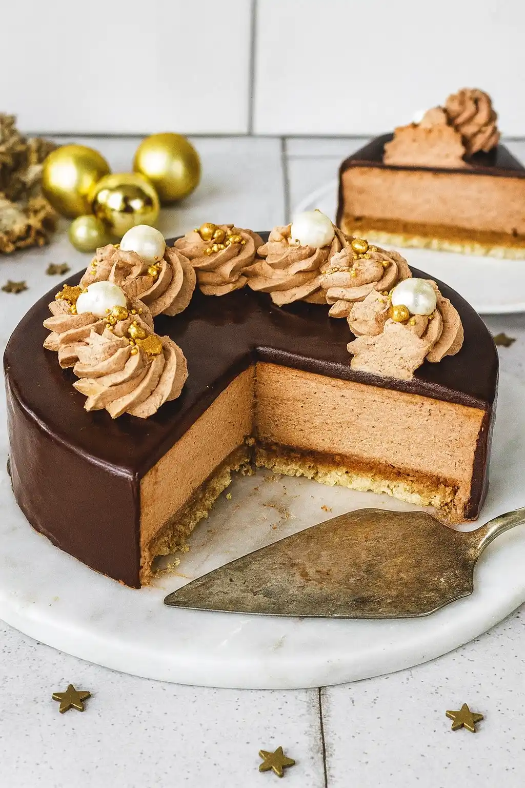 Un somptueux gâteau Trianon rond, orné de crème au chocolat, de perles décoratives et d'un glaçage chocolat brillant, est présenté sur un plateau en marbre blanc. Une tranche a été coupée, révélant les différentes couches : biscuit, croustillant praliné et mousse au chocolat. Des boules dorées et des étoiles décoratives ajoutent une touche festive. Un autre morceau de gâteau Trianon est visible à l'arrière-plan.